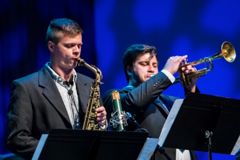 A saxophonist and a trumpeter perform into a microphone while their sheet music sits before them.